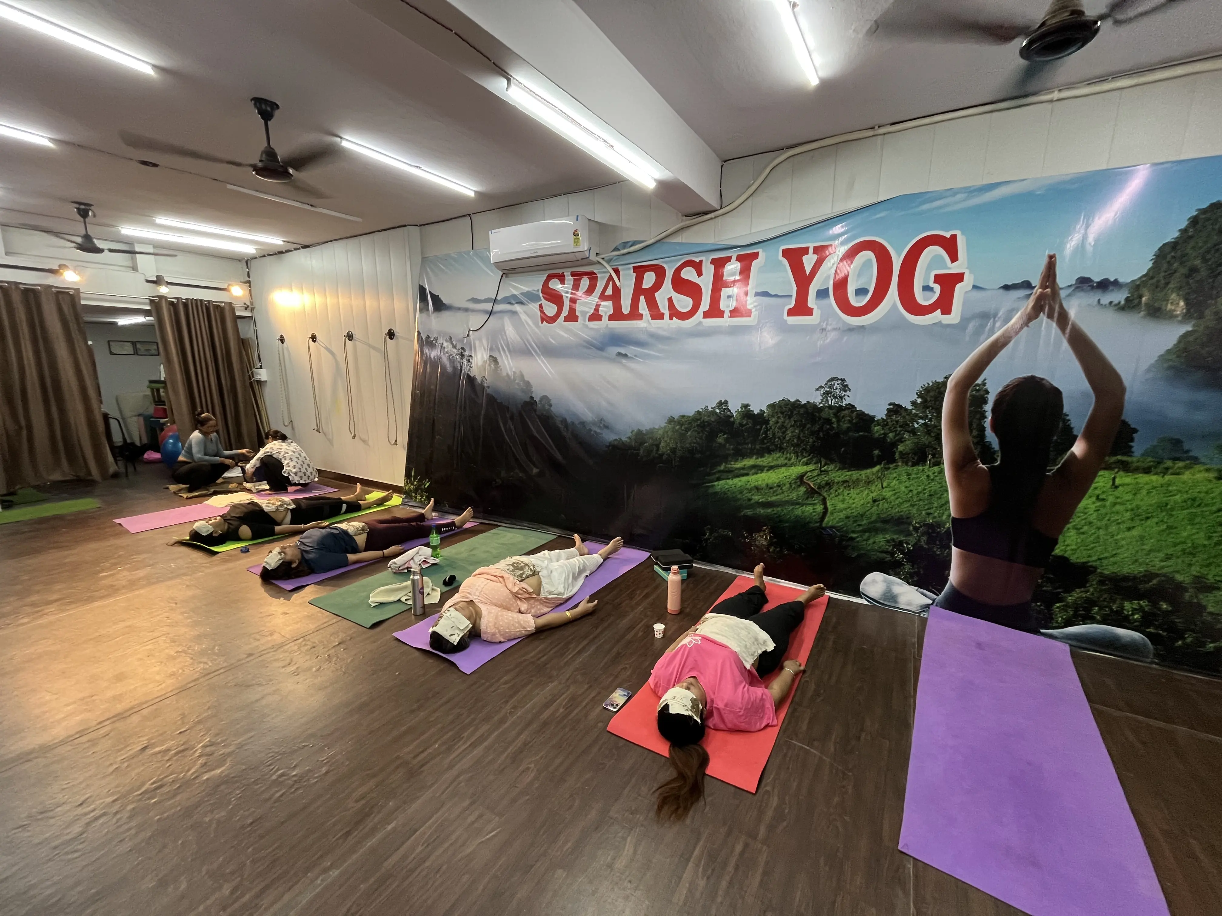 Group mud therapy Mitti Chikitsa session at Sparsh Yog studio Delhi — multiple students resting with mud packs, Sparsh Yog banner visible, guided by Jyoti Singh Gold Medalist